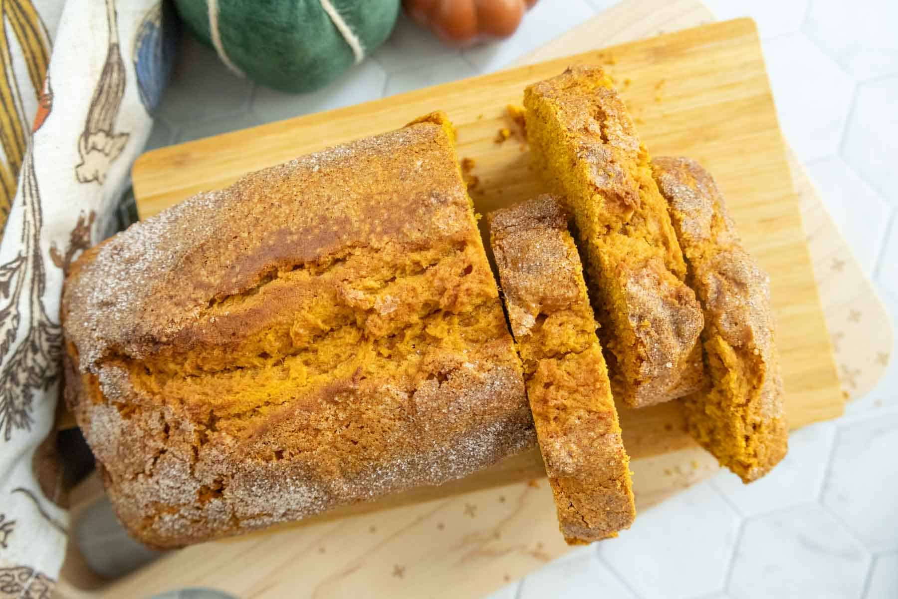 A loaf of pumpkin bread with a sugary crust, partially sliced on a wooden cutting board.