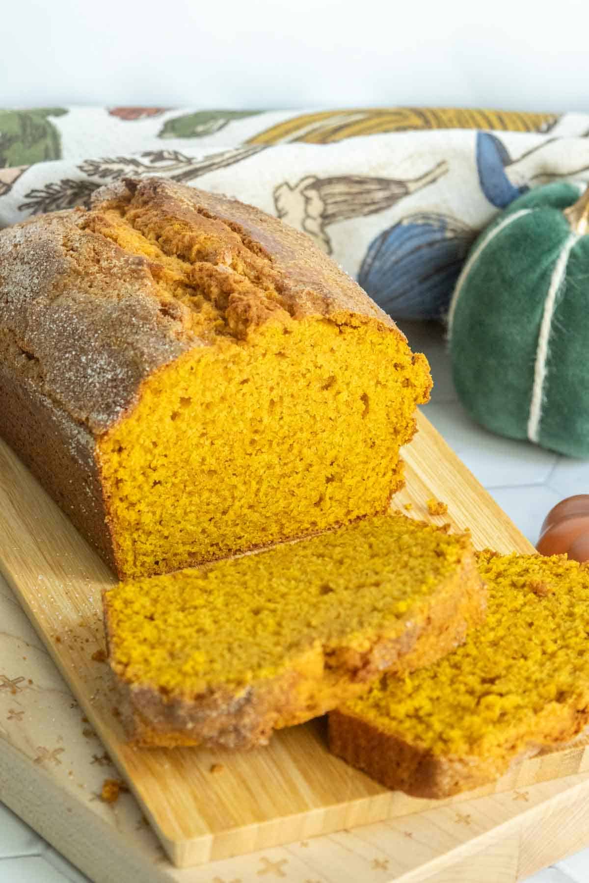 A loaf of pumpkin bread with two slices cut, displayed on a wooden cutting board. A decorative fabric and green pumpkin ornament are in the background.