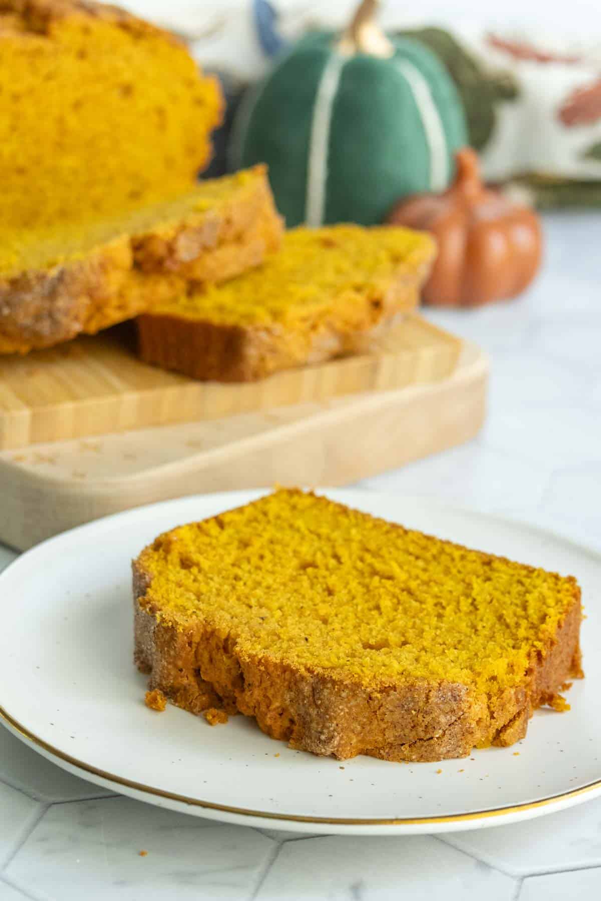 A slice of pumpkin bread on a white plate sits in front of a loaf and two additional slices on a wooden cutting board, with decorative pumpkins in the background.