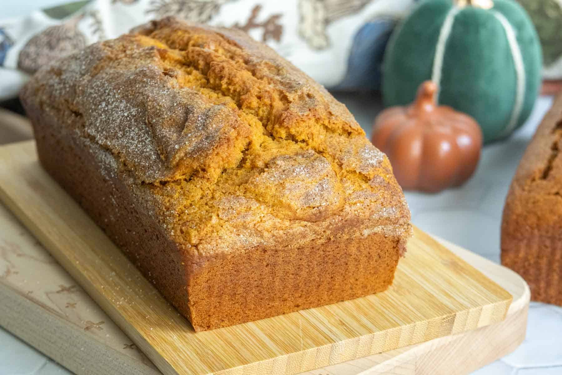 A loaf of pumpkin bread sits on a wooden cutting board, with a small ceramic pumpkin and a decorative cloth in the background.