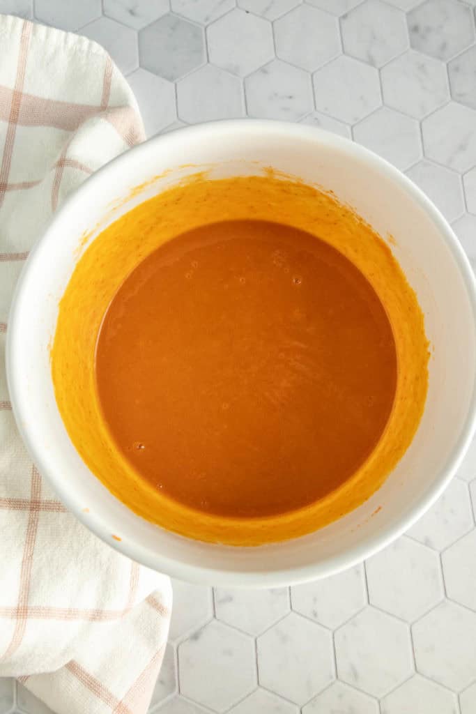 A white bowl filled with orange batter sits on a hexagonal tile countertop next to a beige and white checkered towel.