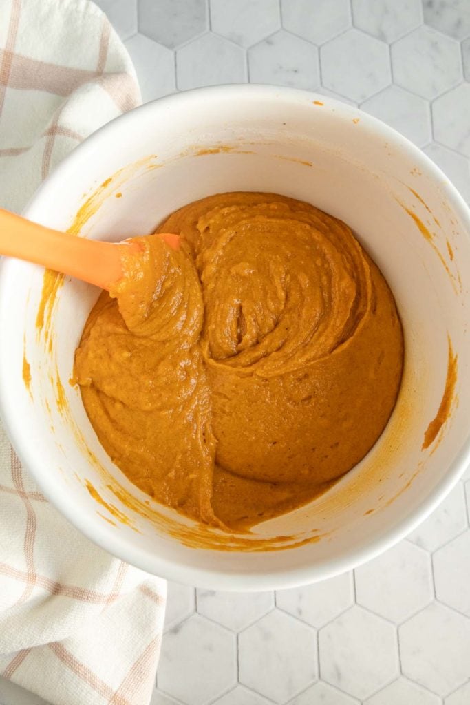 A white mixing bowl filled with thick orange batter and an orange spatula, placed on a hexagonal tile countertop next to a striped cloth.