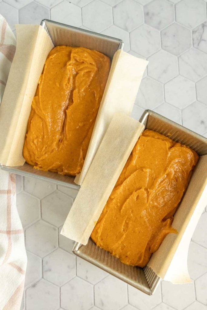 Two loaf pans lined with parchment paper, each filled with an orange batter, are placed on a white hexagonal tile countertop.