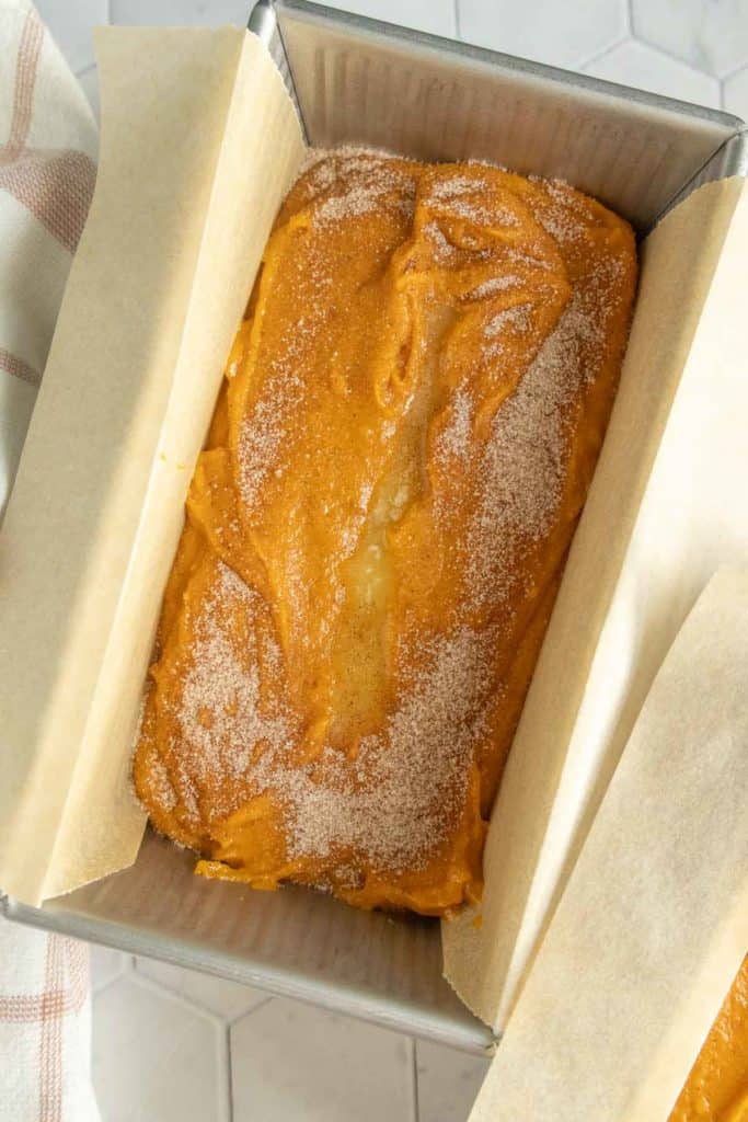 A loaf pan lined with parchment paper holds unbaked pumpkin bread batter topped with a sprinkle of cinnamon sugar.