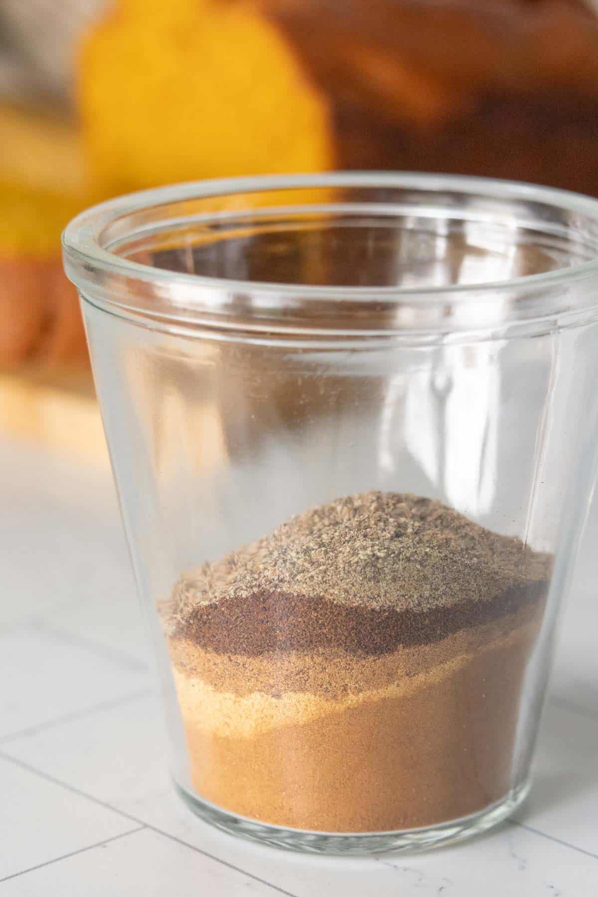 A clear glass jar containing layered ground spices, including cinnamon, ginger, and cloves, on a white surface with blurred bread in the background.