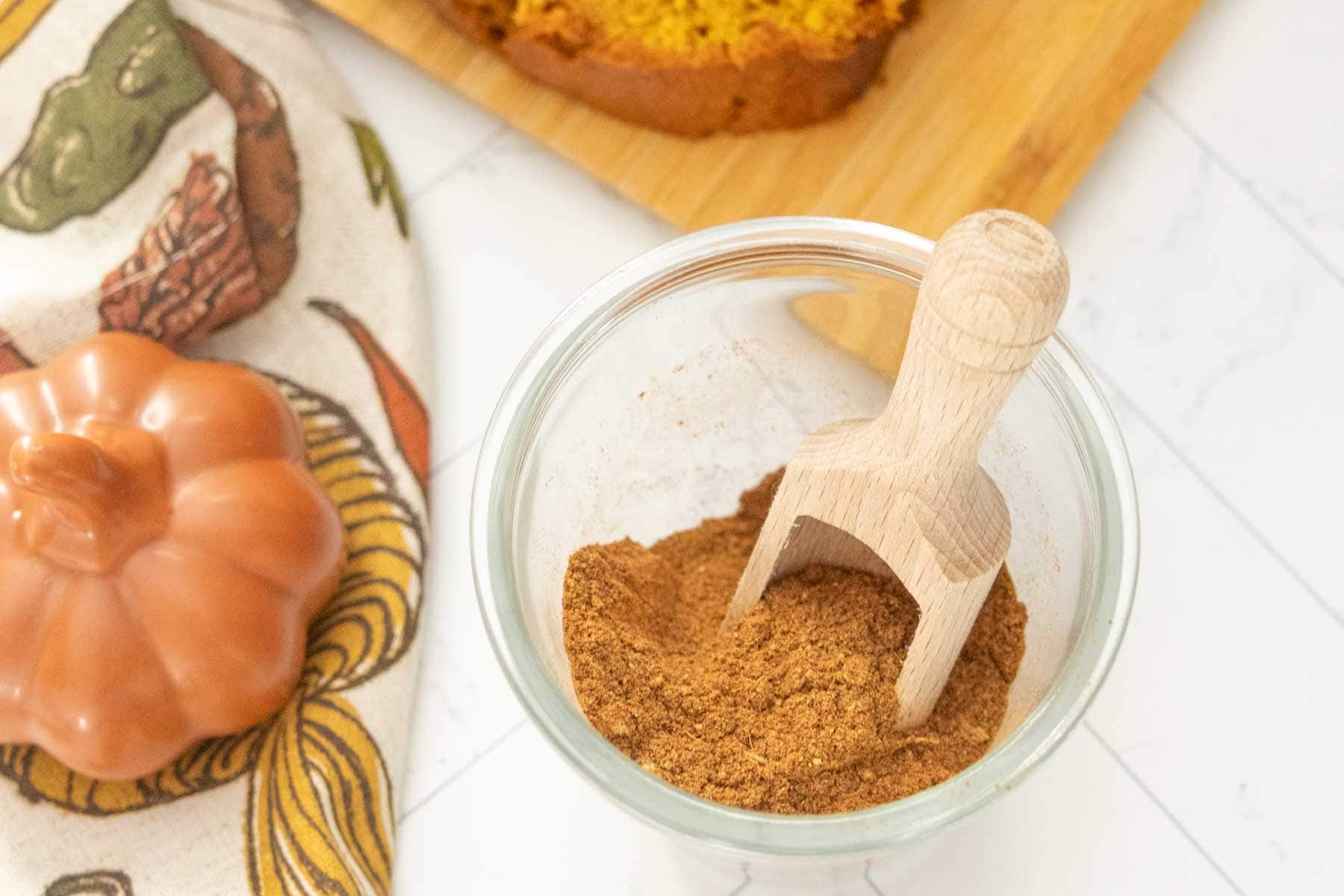 A glass jar filled with ground spice and a wooden scoop, next to a ceramic pumpkin, a patterned cloth, and a loaf of bread on a wooden board.