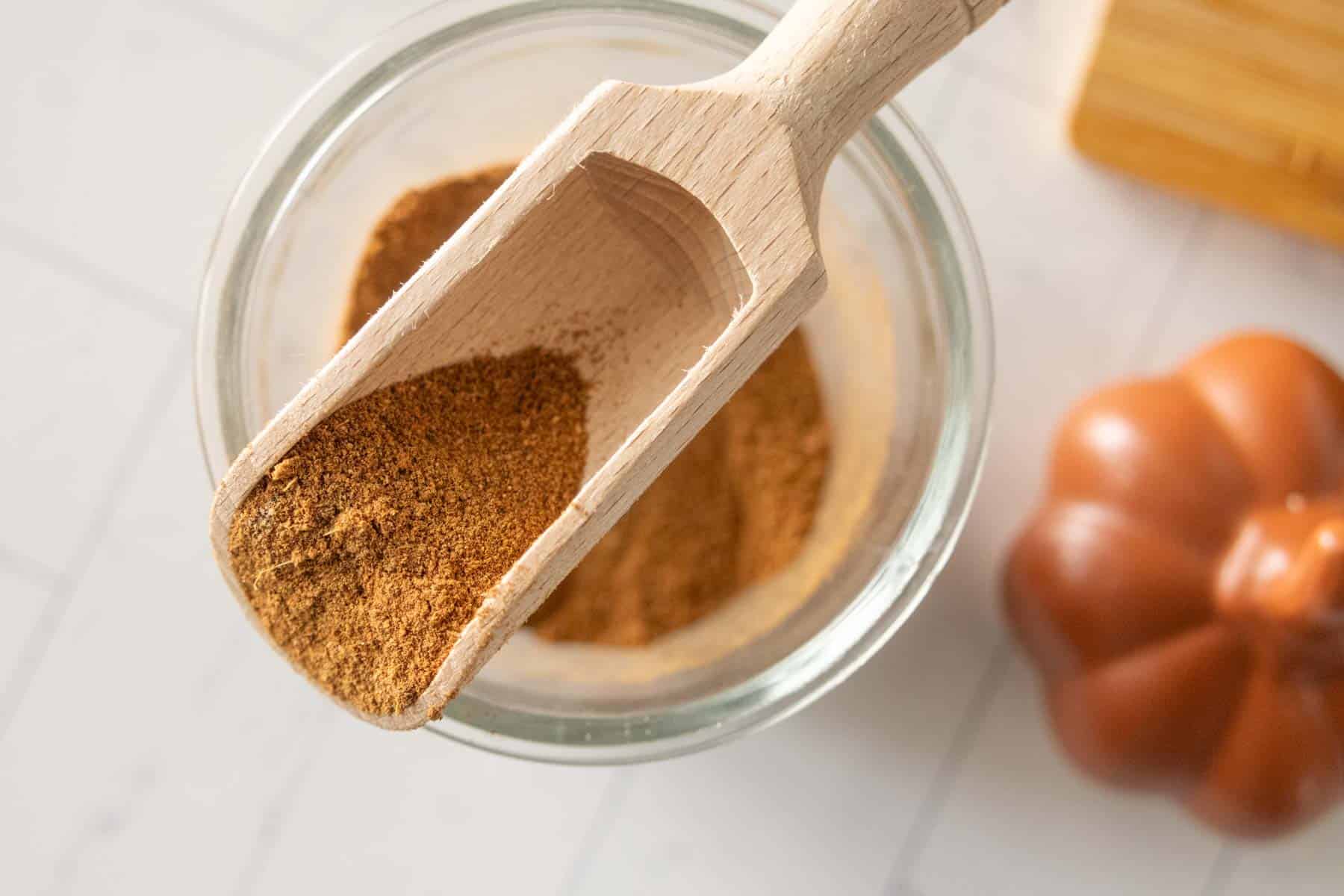 A wooden scoop filled with ground spice rests over a glass jar containing more of the same spice, with a small pumpkin nearby on a white surface.