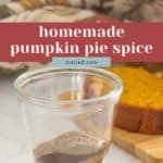 A glass jar filled with layered homemade Pumpkin Pie Spice sits on a counter, with a pumpkin loaf and folded kitchen towels in the background.