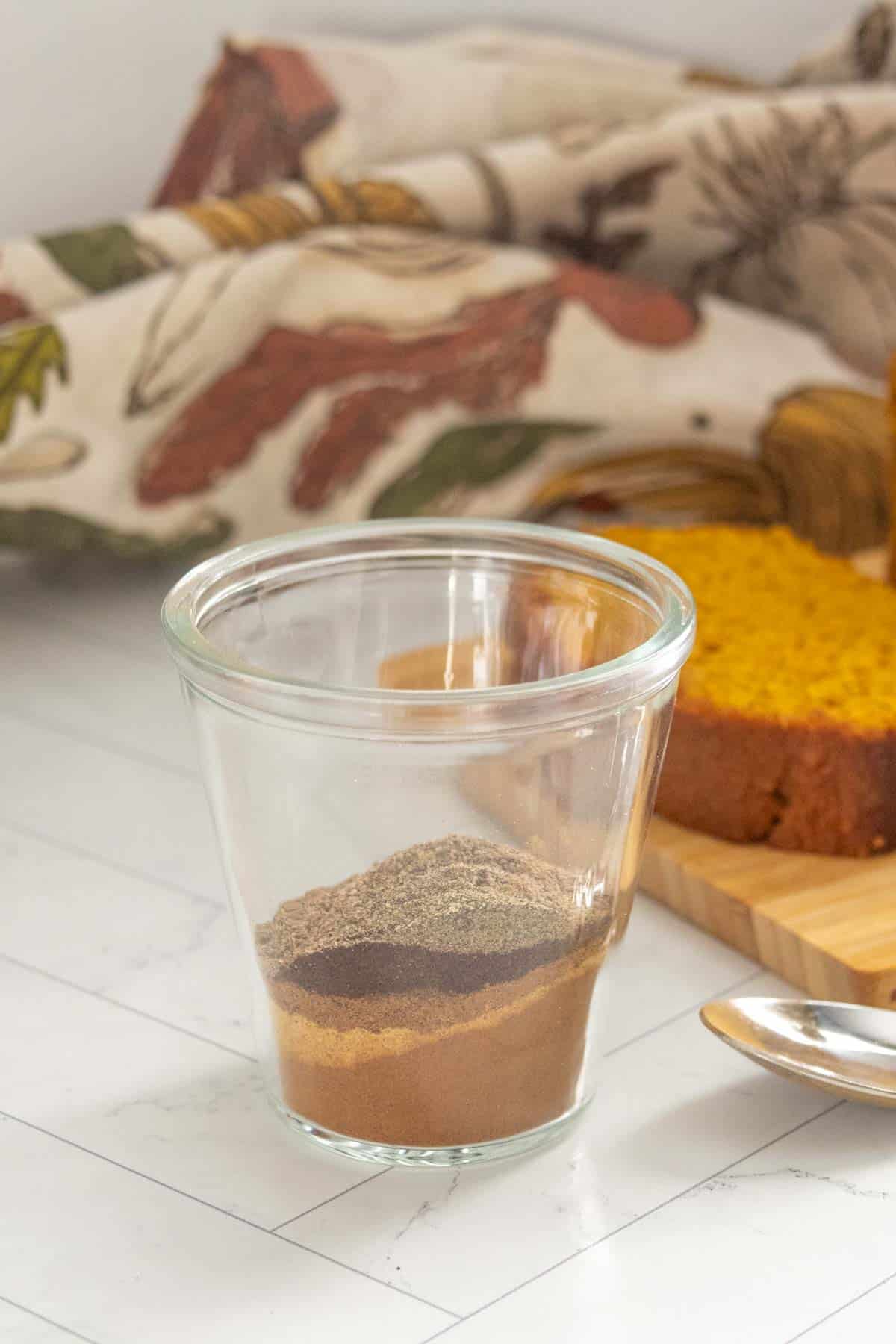 A glass cup contains measured layers of powdered spices, with a spoon nearby and a sliced loaf of bread in the background.