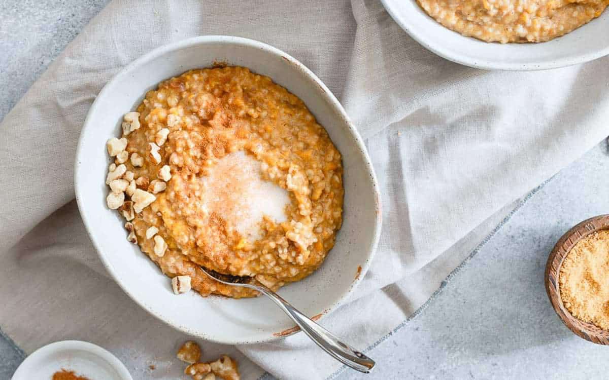 A bowl of oatmeal topped with chopped nuts, brown sugar, and a spoon, placed on a light-colored cloth with another bowl and small dishes nearby.
