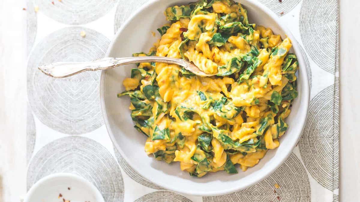 A bowl of pasta mixed with a creamy yellow sauce and green leafy vegetables, with a fork resting inside the bowl.
