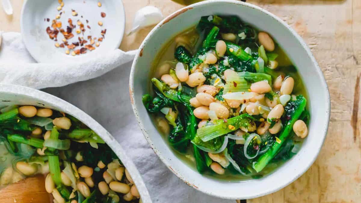 A bowl of cooked greens and white beans in broth, topped with red pepper flakes, sits on a light-colored table next to a small dish of pepper flakes.