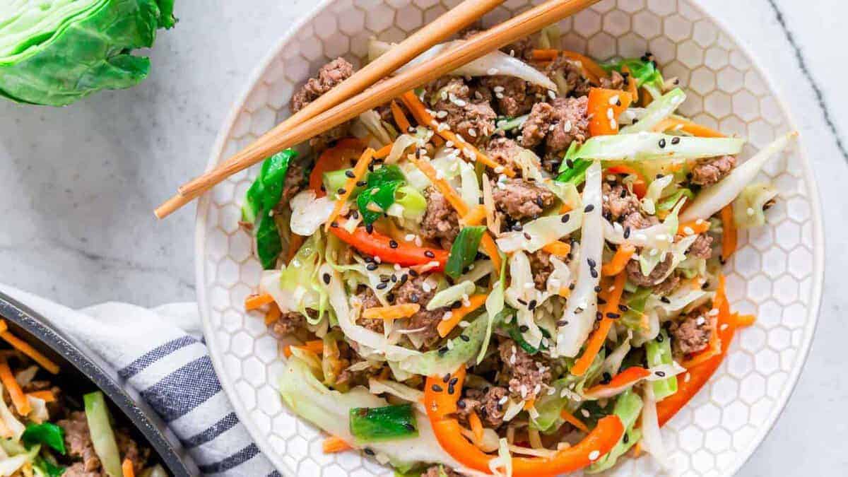 A bowl of stir-fried vegetables, ground meat, and sesame seeds garnished with black and white sesame seeds, with chopsticks resting on the bowl.