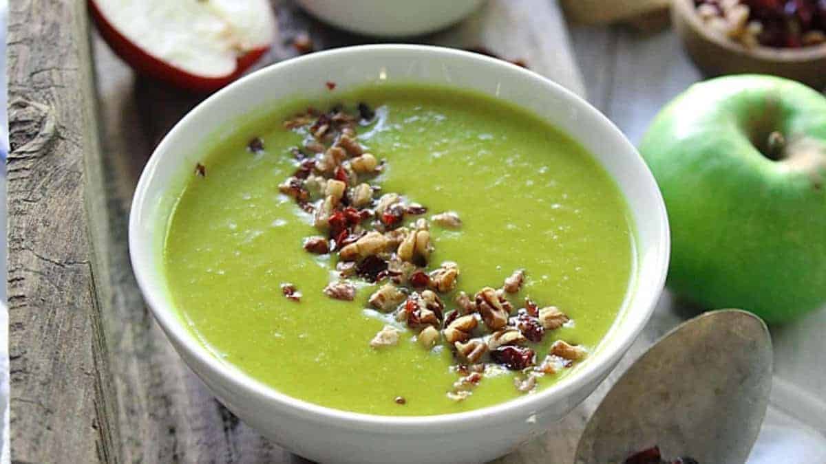 A bowl of green soup garnished with chopped nuts and dried fruit sits on a wooden surface next to a green apple and a spoon.