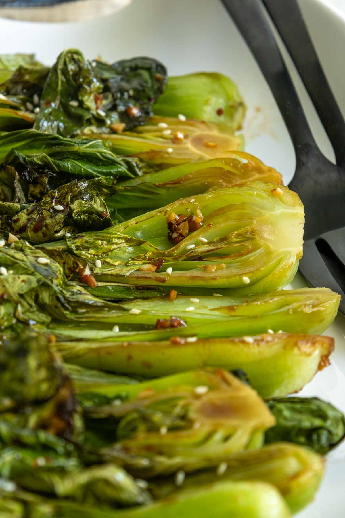 Close-up of roasted baby bok choy halves on a white plate, garnished with sesame seeds and a soy-based sauce, with black serving utensils in the background.
