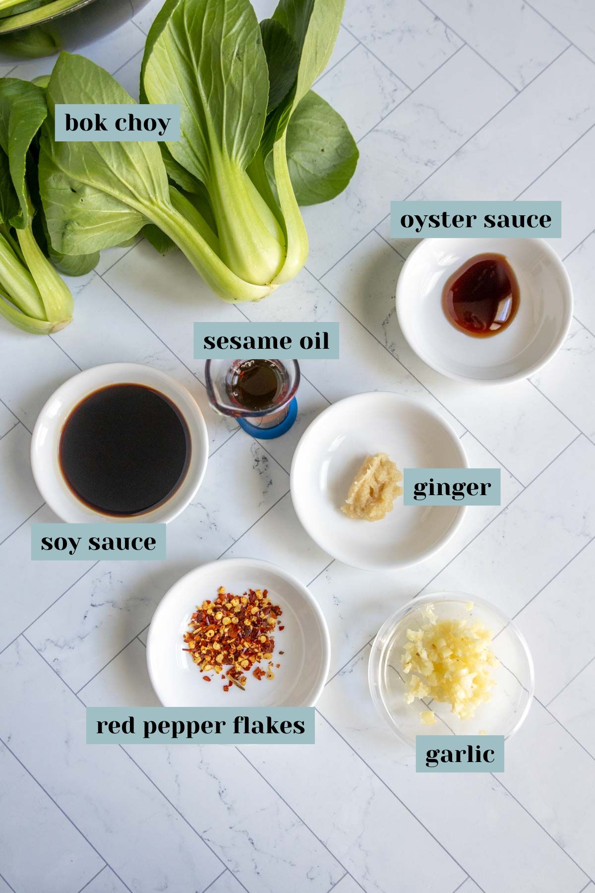 Top-down view of bok choy, soy sauce, sesame oil, oyster sauce, ginger, red pepper flakes, and garlic arranged in bowls on a white surface, each labeled.
