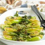 Grilled bok choy halves topped with sesame seeds arranged on a white serving platter with black serving utensils.