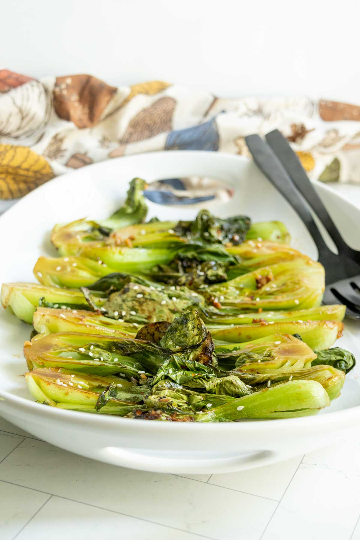 Grilled bok choy halves topped with sesame seeds arranged on a white serving platter with black serving utensils.