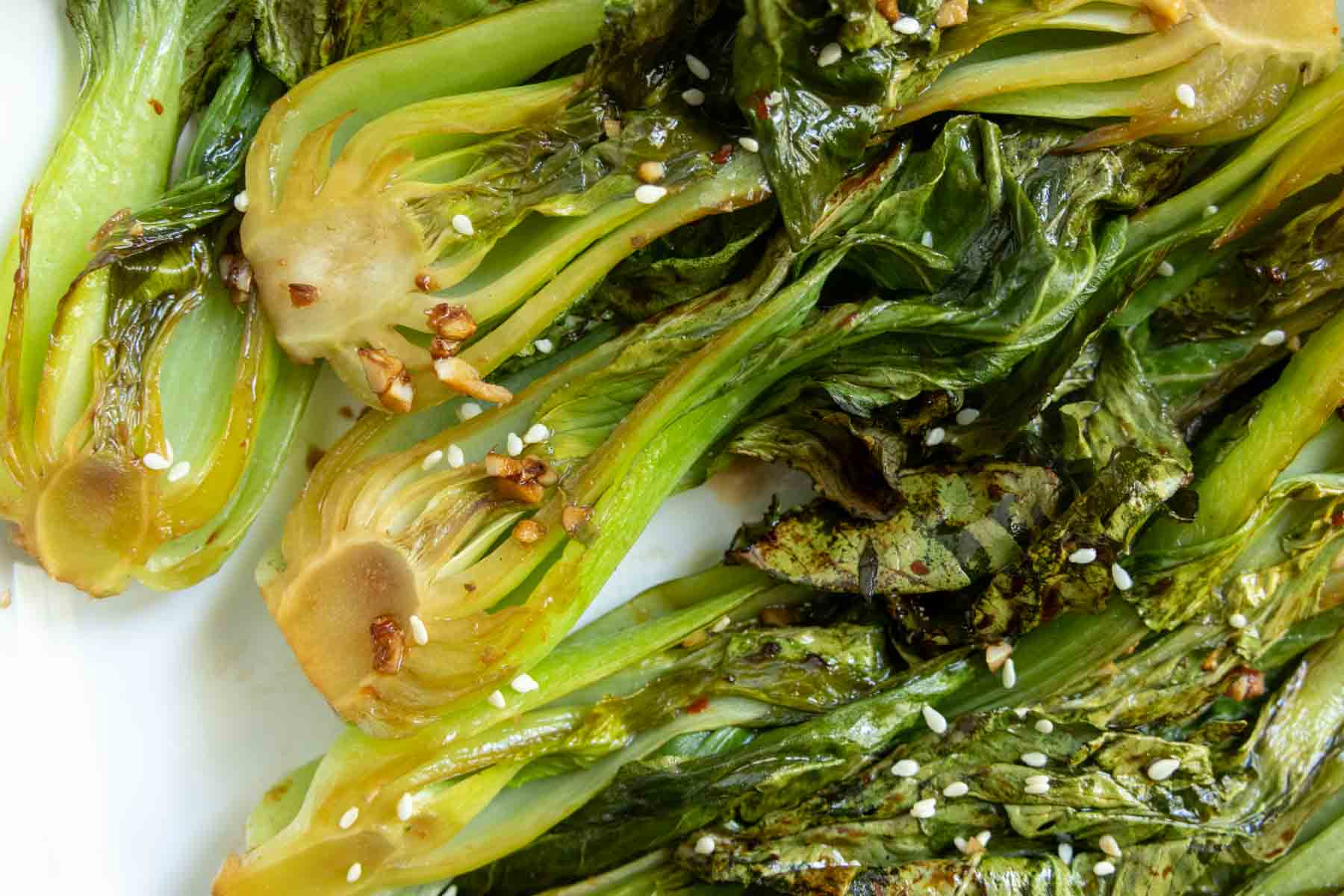 Close-up of roasted bok choy halves garnished with sesame seeds and minced garlic on a white surface.