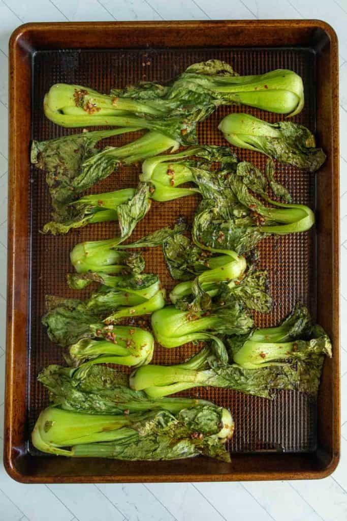 Roasted bok choy halves with seasoning on a dark baking sheet, placed on a light-colored surface.