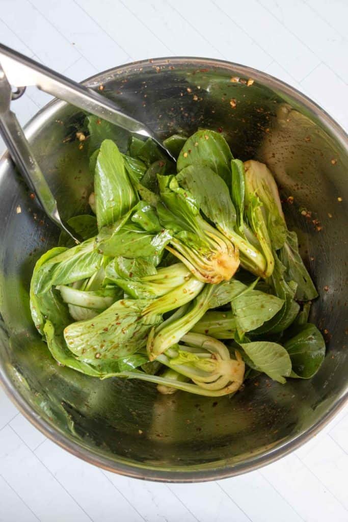 Metal bowl of fresh baby bok choy with seasonings, with metal tongs resting on the edge, placed on a white surface.