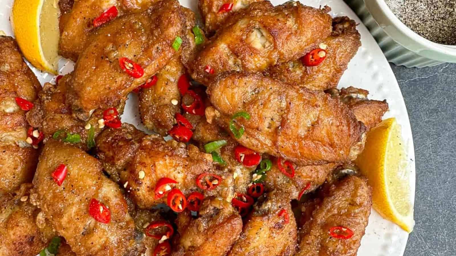 A plate of crispy fried chicken wings garnished with sliced red chilies and green onions, served with lemon wedges on the side.