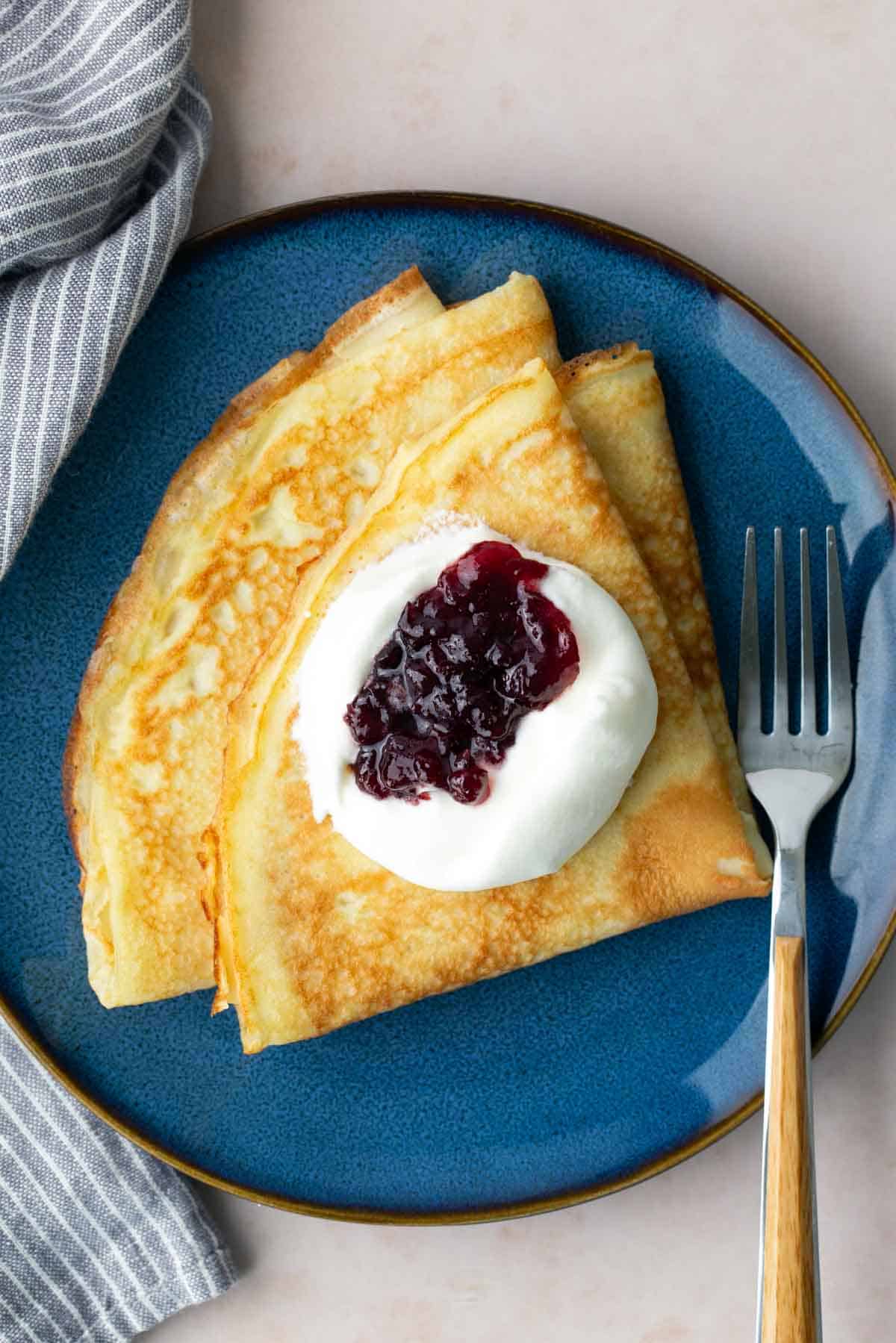 Two folded crepes on a blue plate, topped with a dollop of whipped cream and berry jam, with a fork placed beside them.