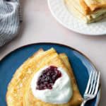 A folded crepe topped with whipped cream and berry jam sits on a blue plate with a fork; a stack of plain crepes is in the background.