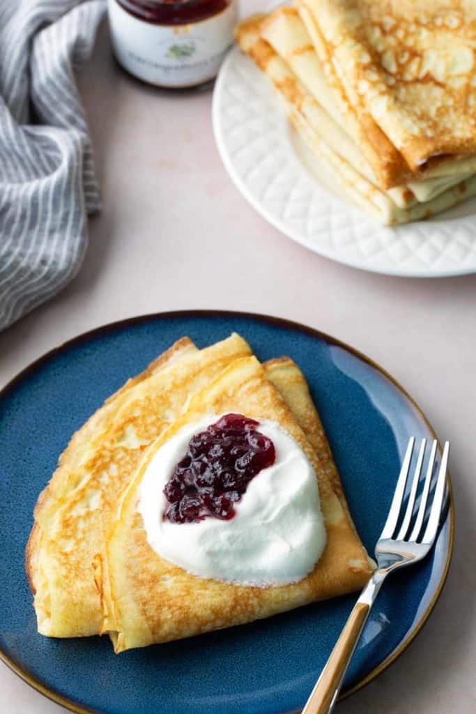 A folded crepe topped with whipped cream and berry jam sits on a blue plate with a fork; a stack of plain crepes is in the background.