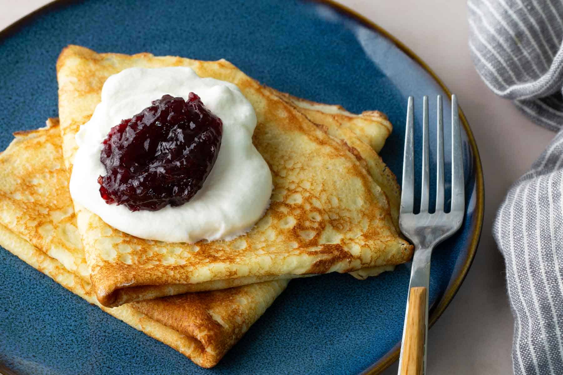 Two folded crepes on a blue plate topped with whipped cream and a dollop of berry jam, with a fork placed beside them.