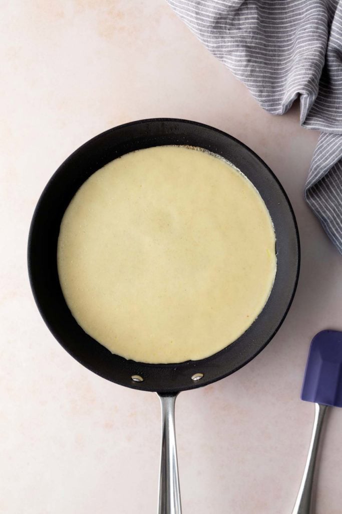 A nonstick frying pan with uncooked crepe batter spread evenly, a striped towel and a purple spatula nearby.