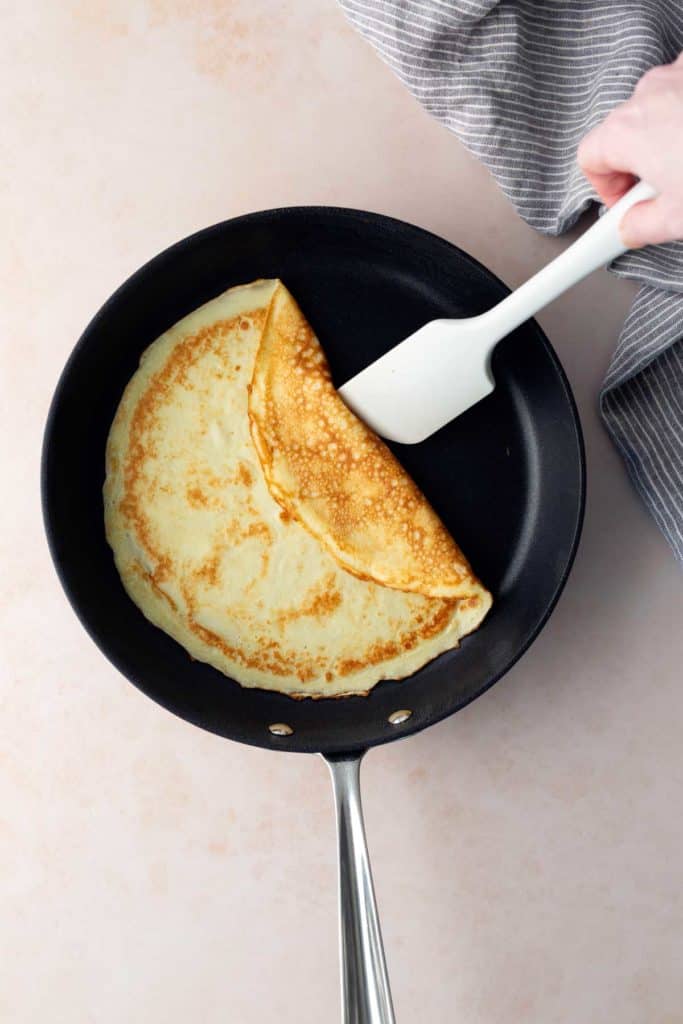 A hand uses a white spatula to fold a crepe in a black frying pan, with a gray striped cloth partially visible on the side.