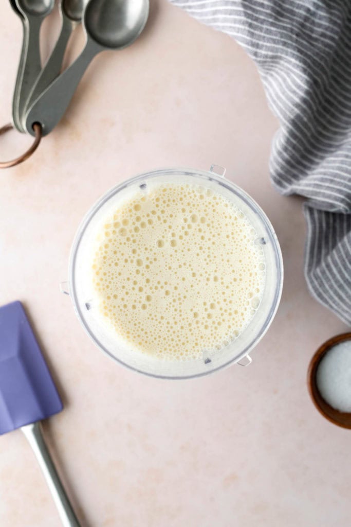 A blender cup filled with frothy liquid, surrounded by measuring spoons, a striped kitchen towel, a purple spatula, and a small bowl of white granulated sugar on a countertop.