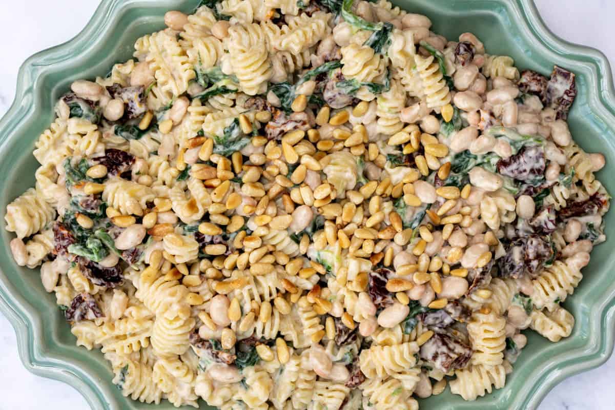 A ceramic dish filled with rotini pasta mixed with white beans, spinach, sun-dried tomatoes, creamy sauce, and topped with toasted pine nuts.