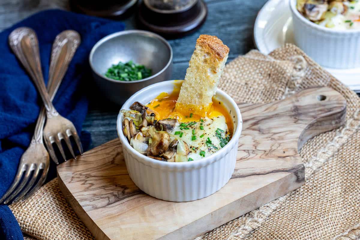 A ramekin with a baked egg dish topped with mushrooms and herbs, served with a slice of toasted bread on a wooden board.