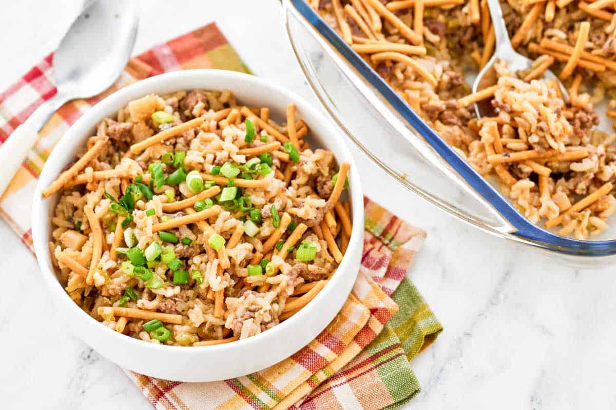 A white bowl and a glass baking dish contain a rice casserole with ground meat, chow mein noodles, and chopped green onions, set on a plaid cloth with a spoon nearby.