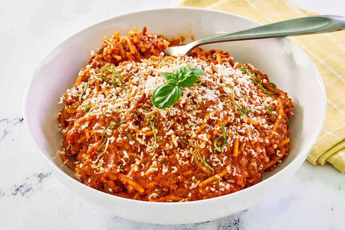 A bowl of spaghetti with red meat sauce, topped with grated cheese and fresh basil, with a spoon resting inside the bowl.