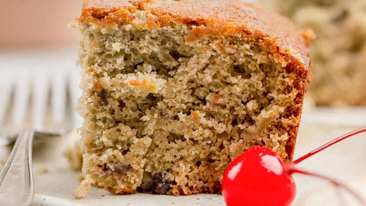 A slice of banana cake on a plate with a fork beside it and a red cherry in the foreground.