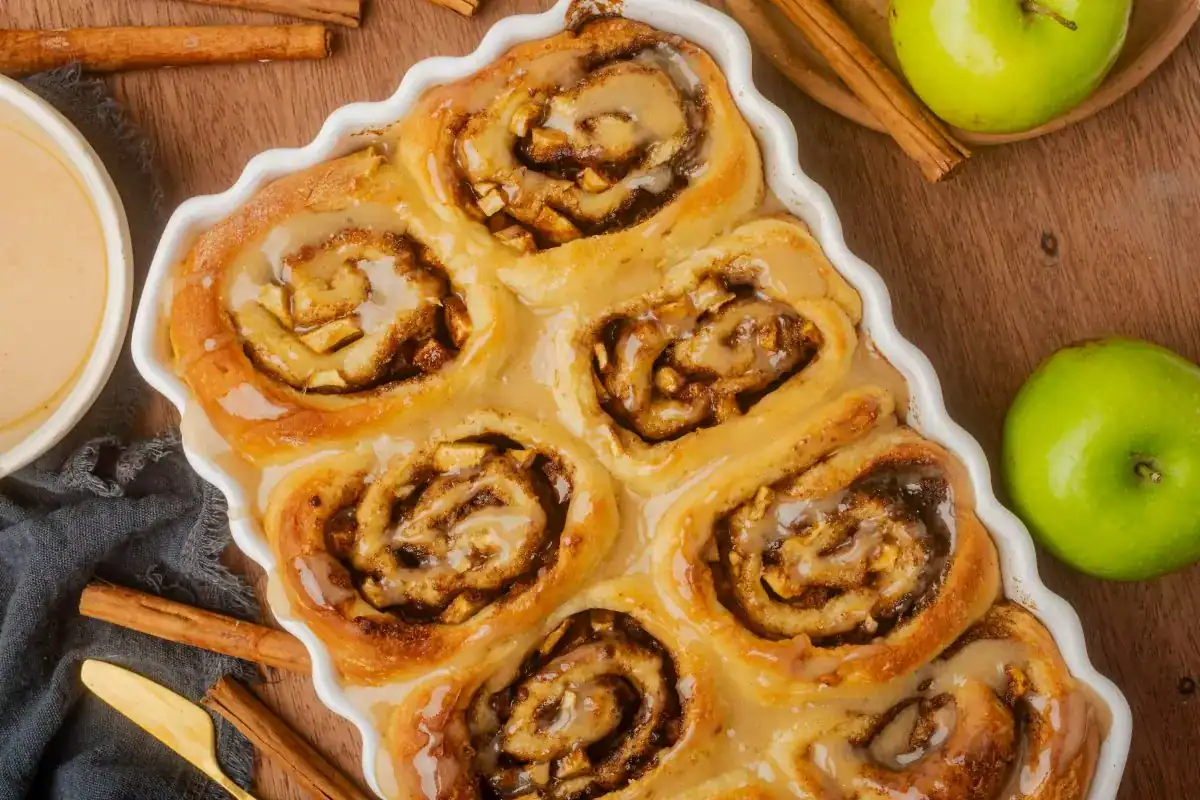 A baking dish with six glazed cinnamon rolls sits on a wooden surface, surrounded by green apples, cinnamon sticks, and a bowl of icing.