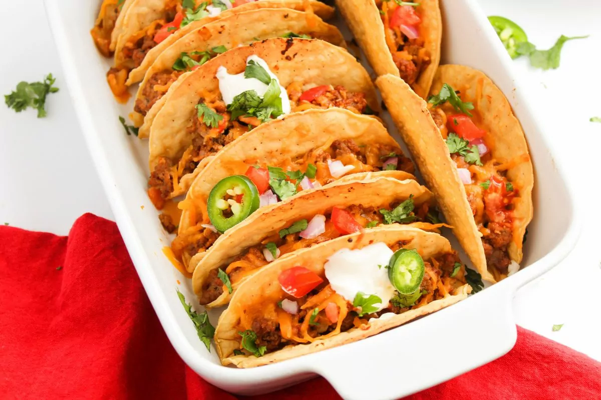 A white baking dish filled with several hard shell tacos topped with chopped tomatoes, jalapeños, onions, cilantro, and sour cream. A red cloth is partially visible beside the dish.