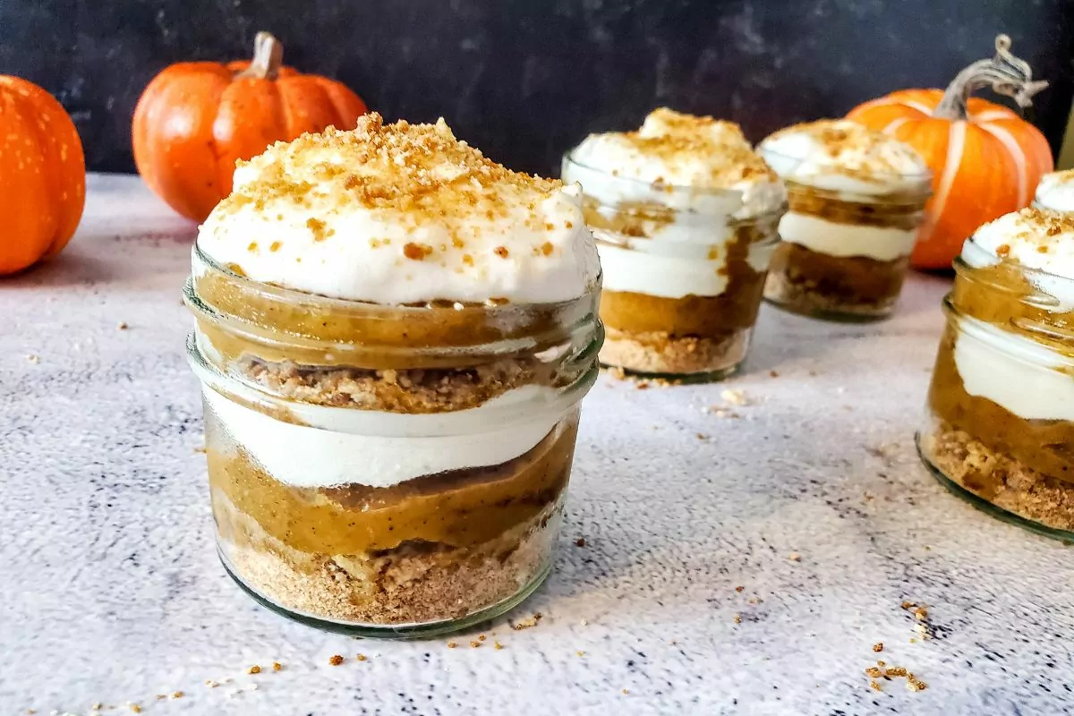 Mini pumpkin parfaits in glass jars with layers of pumpkin filling, whipped cream, and crumb crust, set on a light surface with small pumpkins in the background.