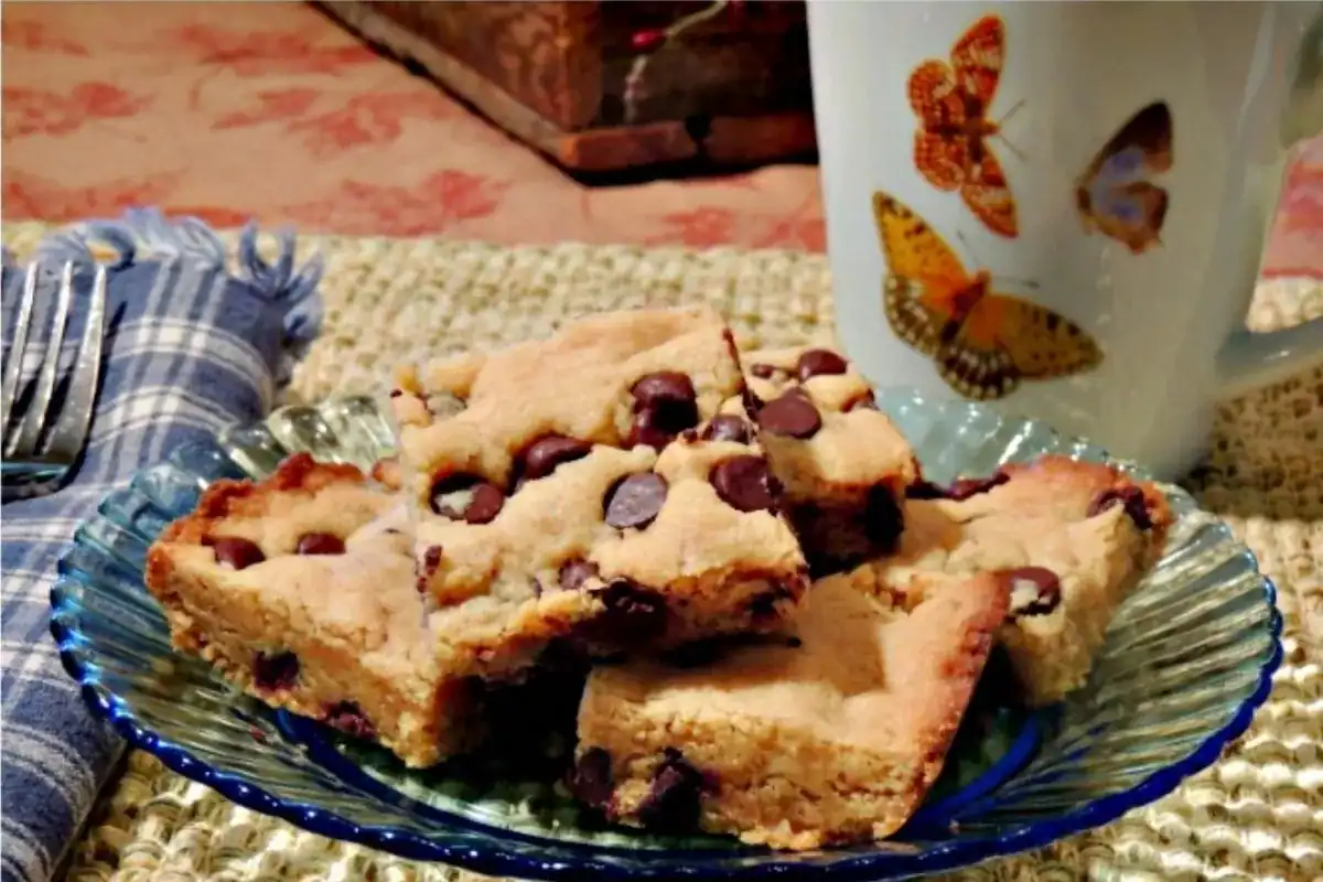 A glass plate with several chocolate chip cookie bars sits on a woven placemat next to a napkin, fork, and a mug decorated with butterfly illustrations.
