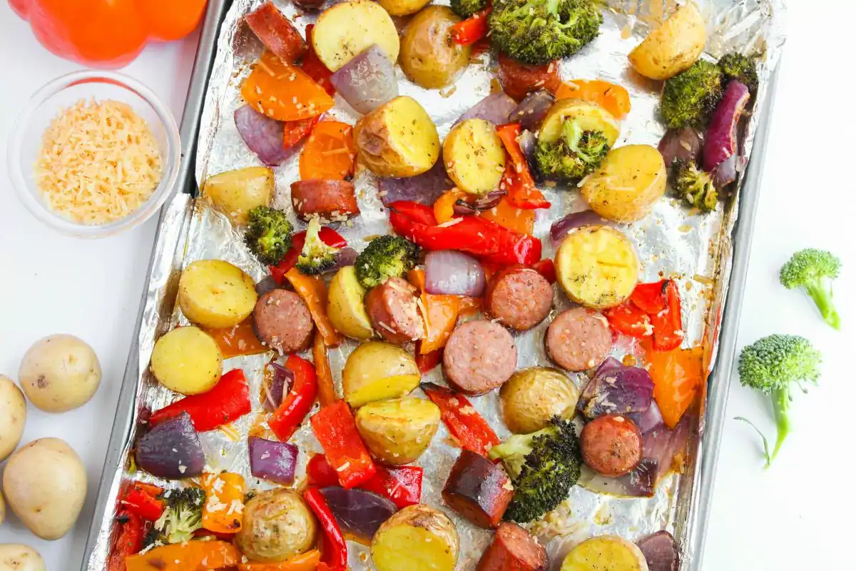 Sheet pan with roasted potatoes, sliced sausage, broccoli, red onion, and red bell pepper on foil. Small bowls of cheese and vegetables are nearby.