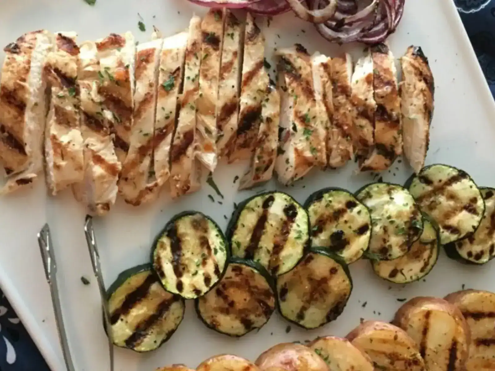 Sliced grilled chicken breast, grilled zucchini rounds, potato slices, and red onion served on a white platter with metal tongs.
