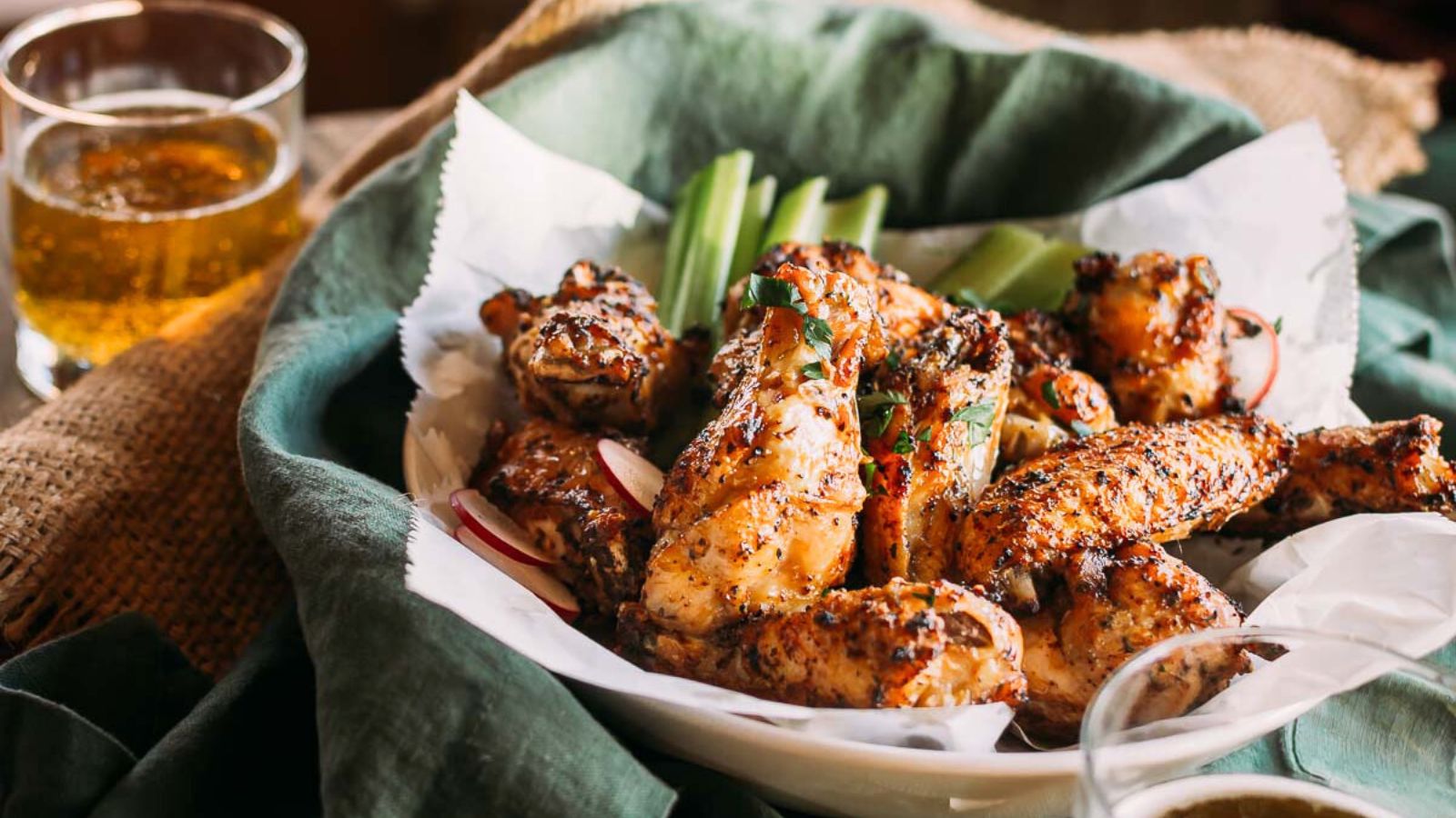 A plate of seasoned grilled chicken wings with celery sticks, served on parchment paper, next to a glass of beer.