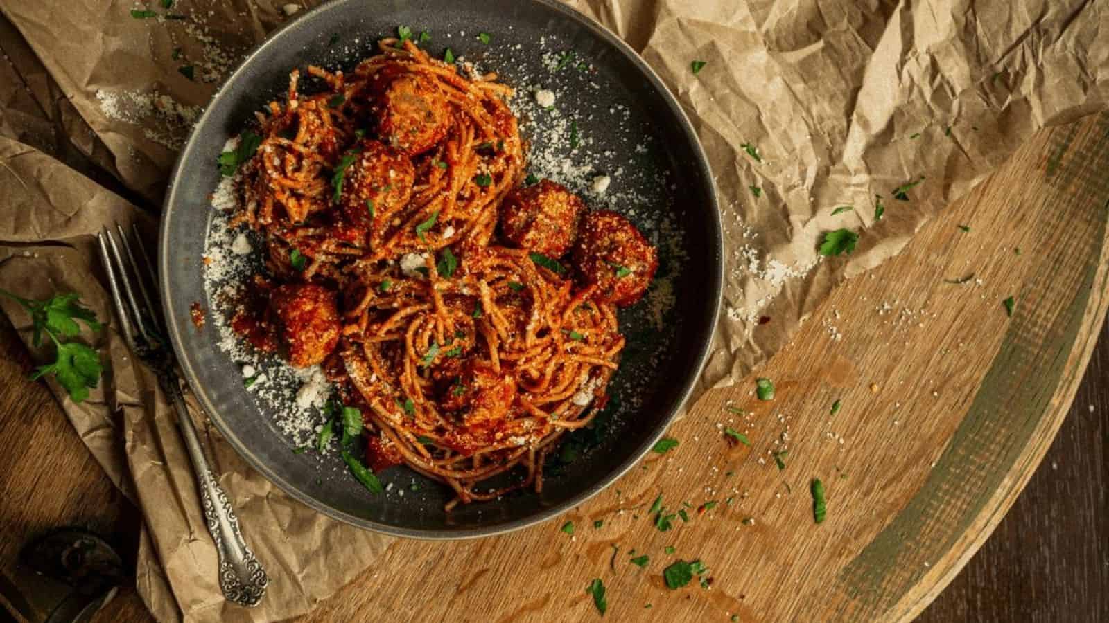 A bowl of spaghetti with meatballs in tomato sauce, garnished with grated cheese and parsley, sits on a crumpled brown paper with a fork and knife beside it.