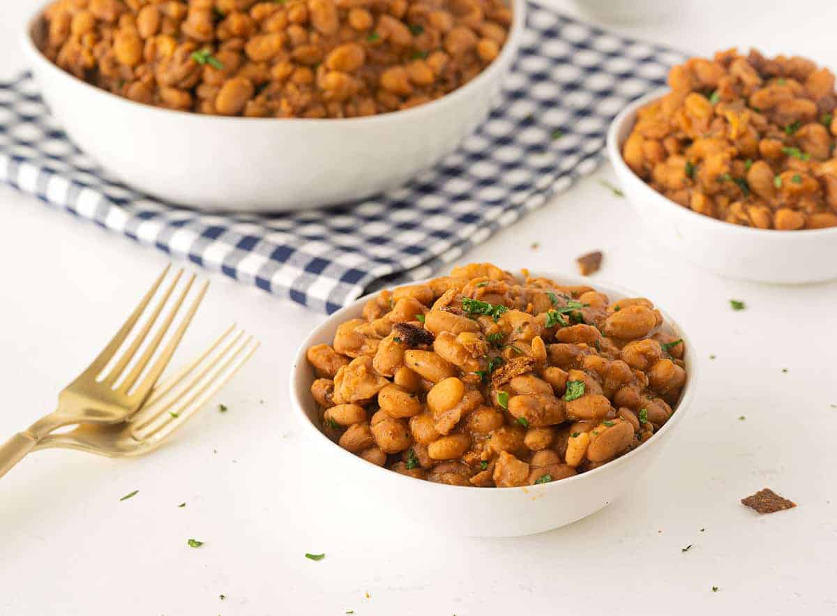 White bowls filled with baked beans garnished with herbs are placed on a white surface next to a gold fork and a blue checkered napkin.