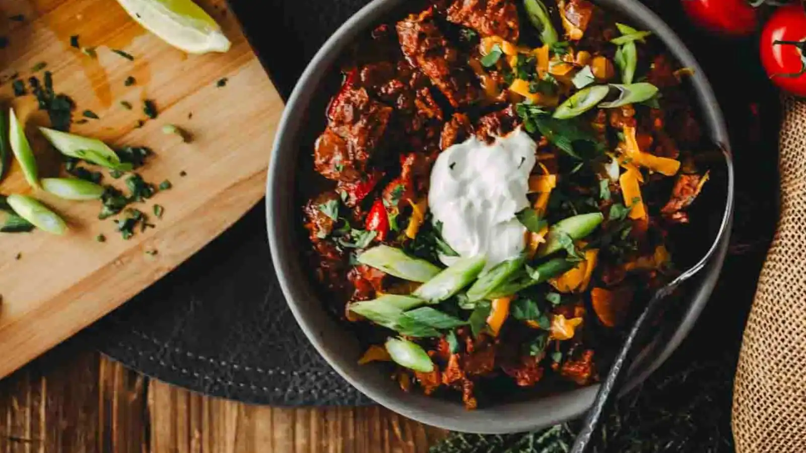 A bowl of chili topped with sour cream, sliced green onions, and shredded cheese sits next to a cutting board with chopped herbs and lime wedges.