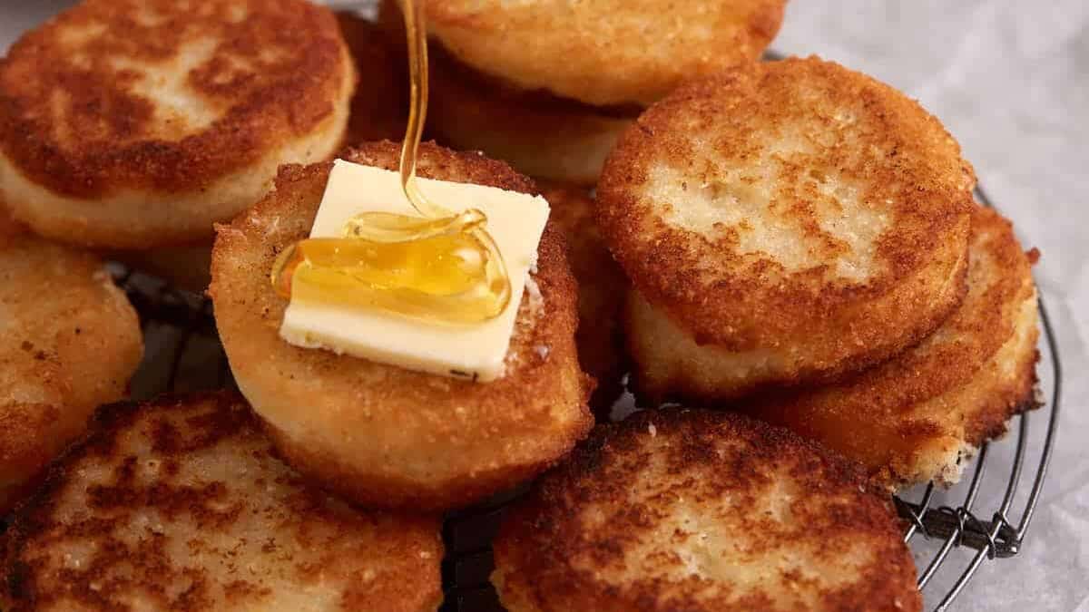 Several golden-brown potato cakes are stacked on a wire rack, with one topped with a pat of butter and honey being drizzled over it.