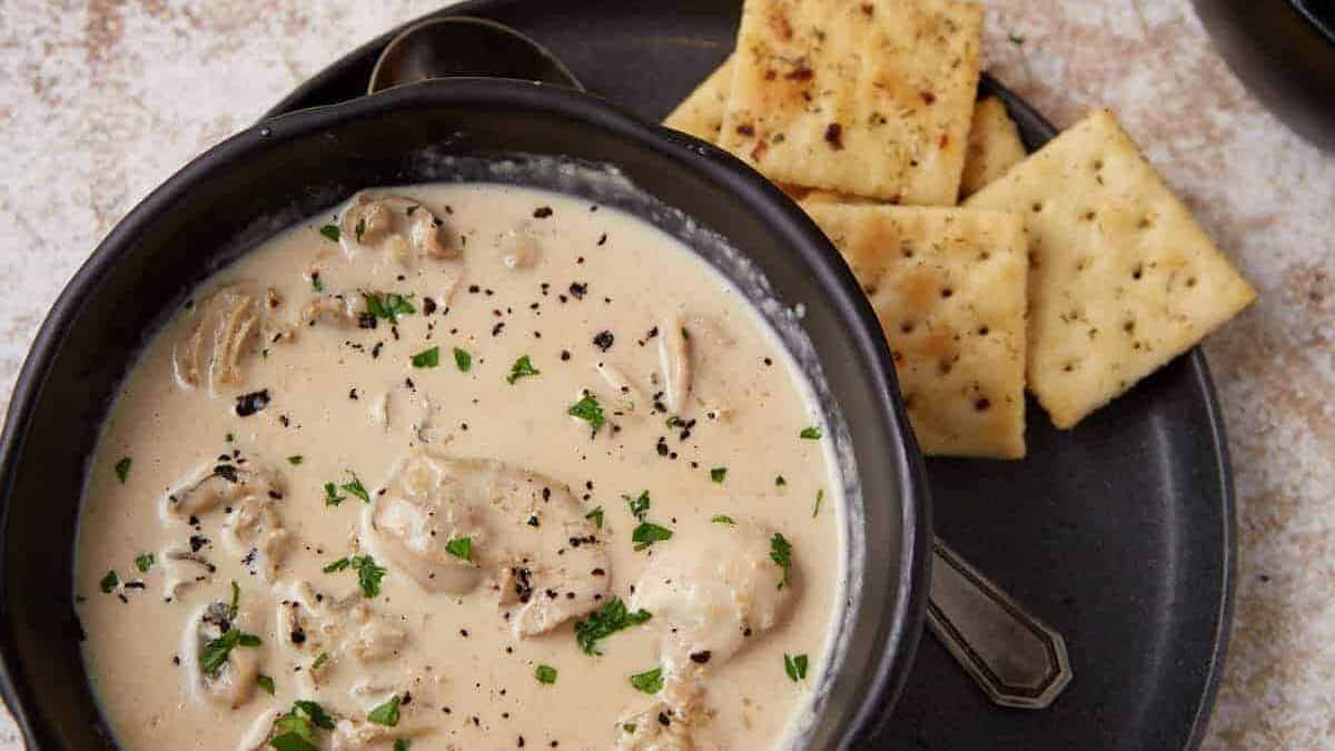 A bowl of creamy clam chowder topped with parsley and black pepper, served with saltine crackers on a dark plate.